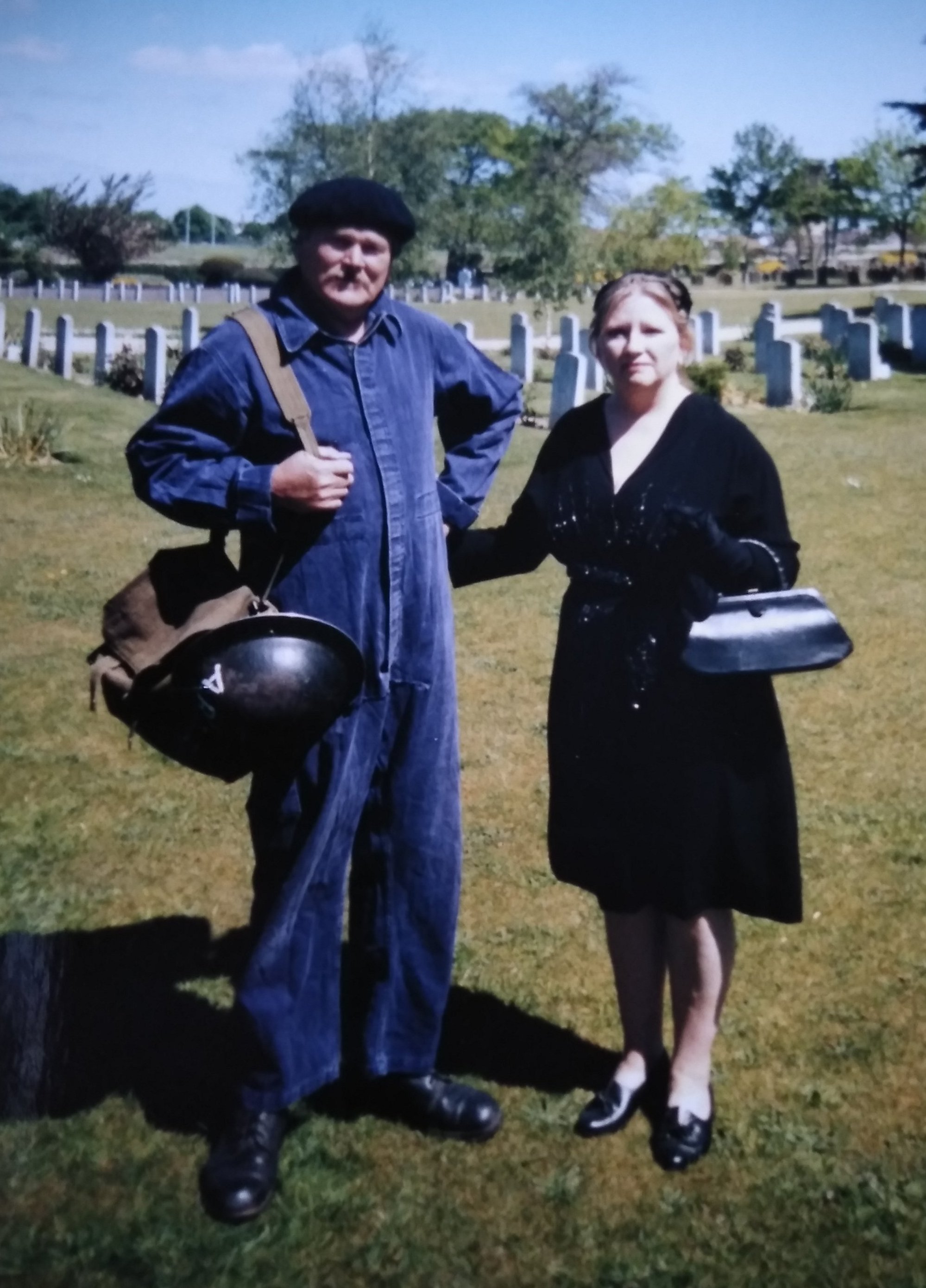 A man and woman dressed in 1940s attire stand in a cemetery.