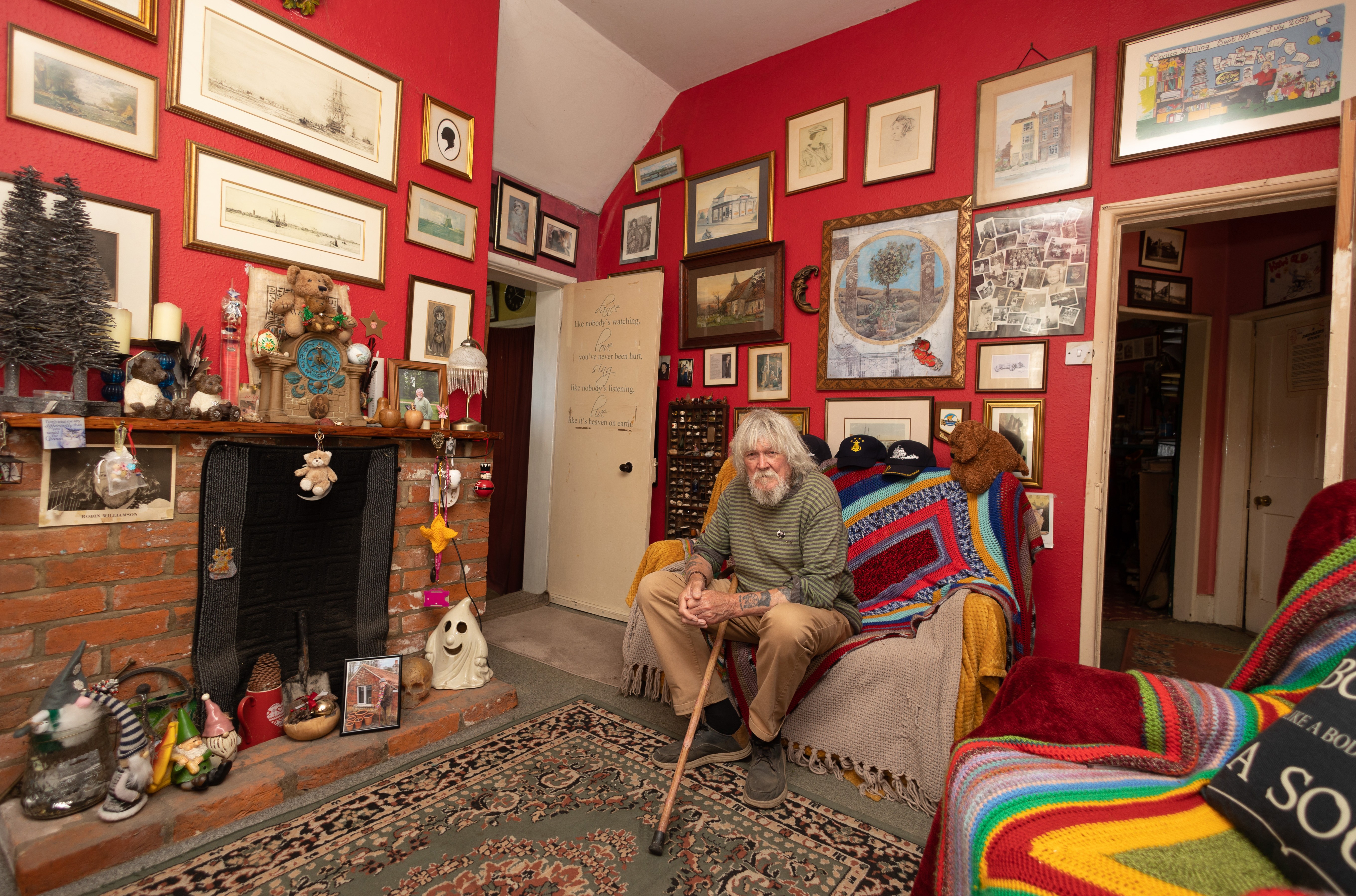 A man sits in a chair in a room filled with framed pictures and other decorations.