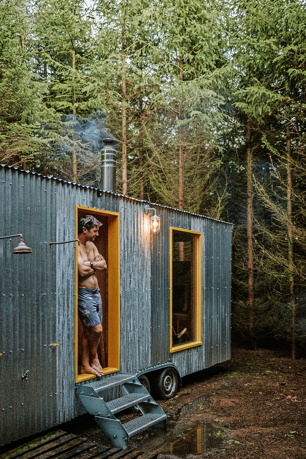 Man standing in doorway of a small metal sauna in the woods.